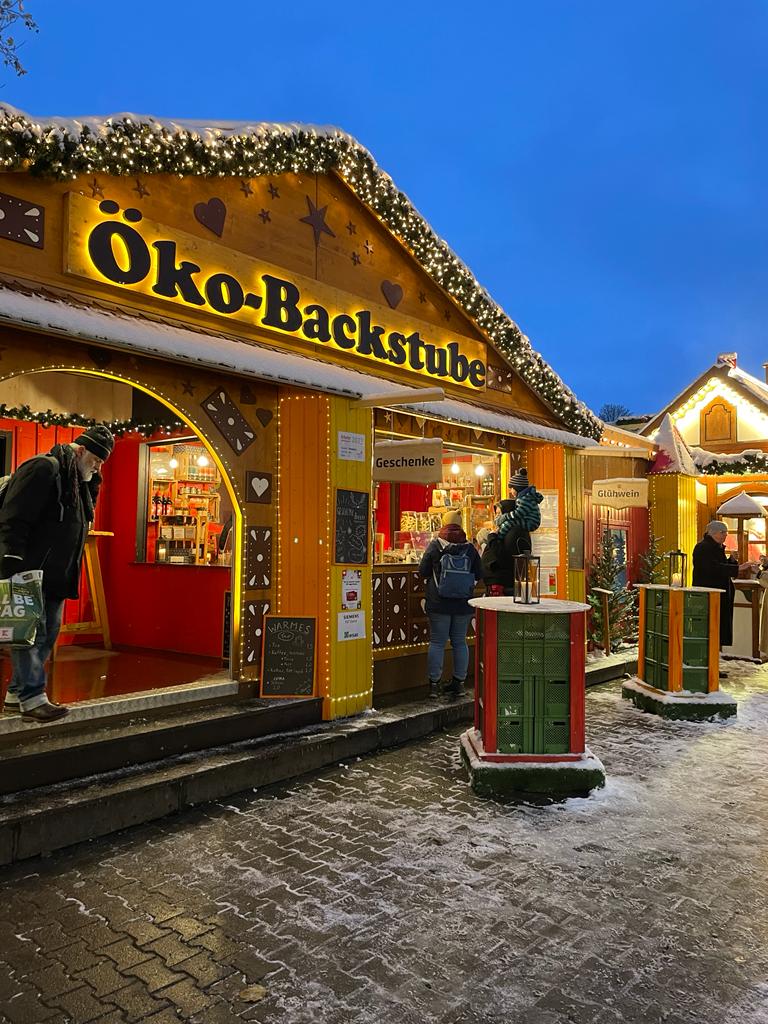 Ansicht Weihnachtsmarktstand von Außen mit Menschen davor und Leuchtschriftzug Oeko-Backstube